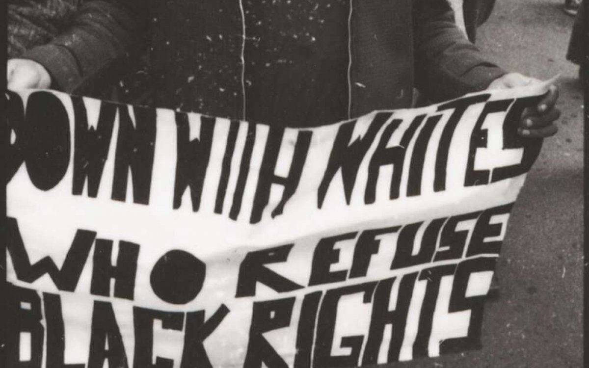 A black and white photo of an Indigenous Australian carrying a sign in a rally of black and white people, that says 'Down with whites who refuse black rights'.