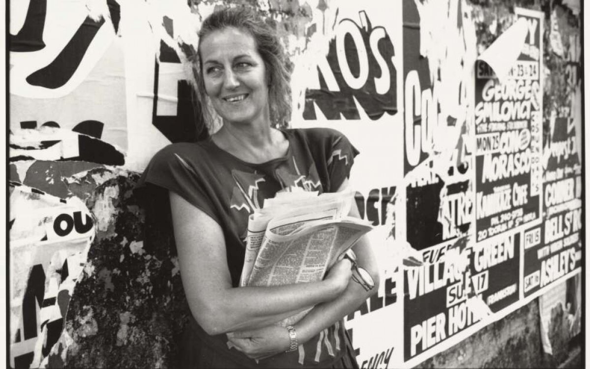A black and white photograph of a woman holding a stack of newspapers. She is smiling and looking off camera. She is standing in front of a wall that is plastered with posters, playbills, advertising and graffiti. At the bottom of the image is a margin. There is handwriting that reads "GERMAIN GREER 1988" and "BRENDAN HENNESSY 89"