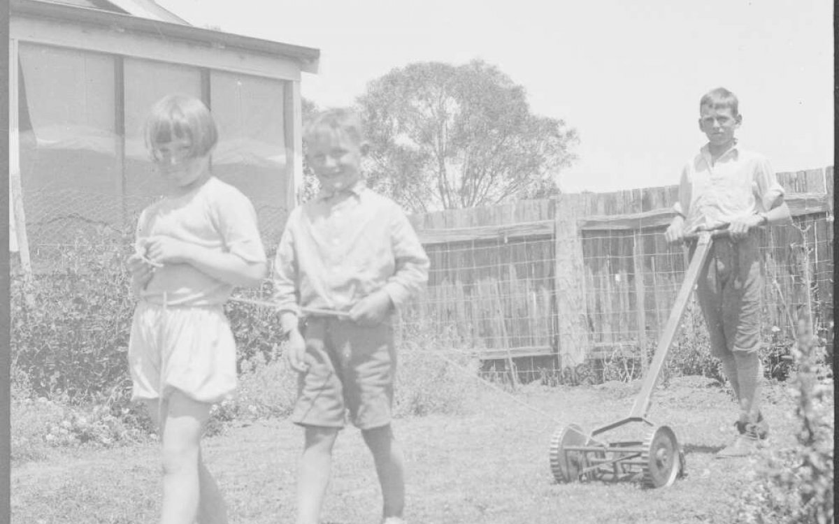 black and white photo of two young boys standing in front of their father mowing the lawn with manual mower