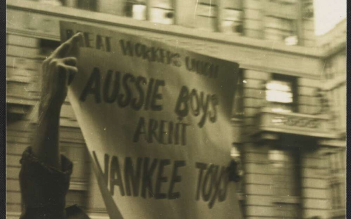 A sepia toned photo of a man in a hat holding a paper sign in a city street, saying 'Meat Workers union Aussie boys aren't Yankee toys'