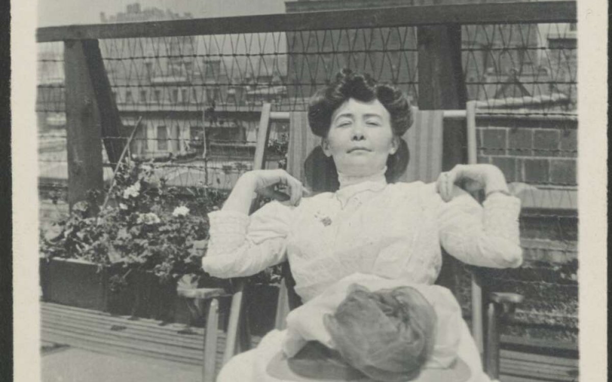 A black and white photograph of a woman reclining in a deck chair. She is wearing a large white dress with a hat sitting on her lap.