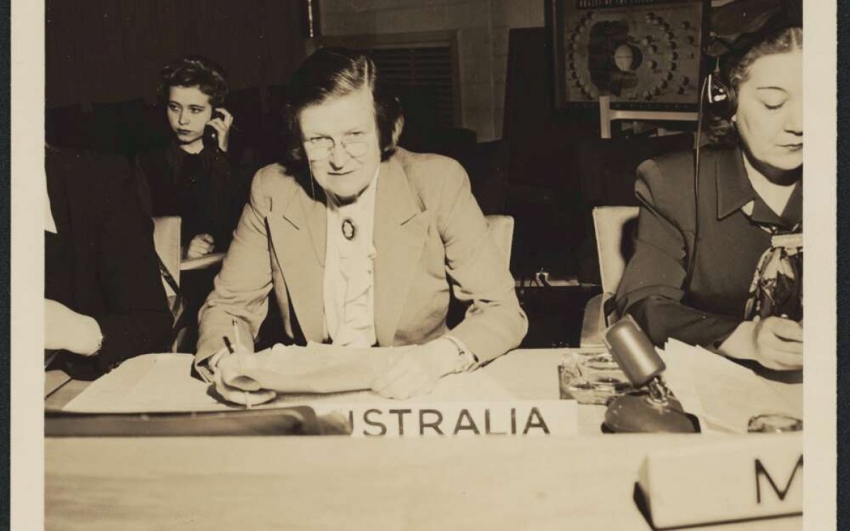 A sepia photo of a woman sitting at a desk. She is sitting infront of a sign that reads "--STRALIA". The first letters are obscured. She is holding a stack of papers.