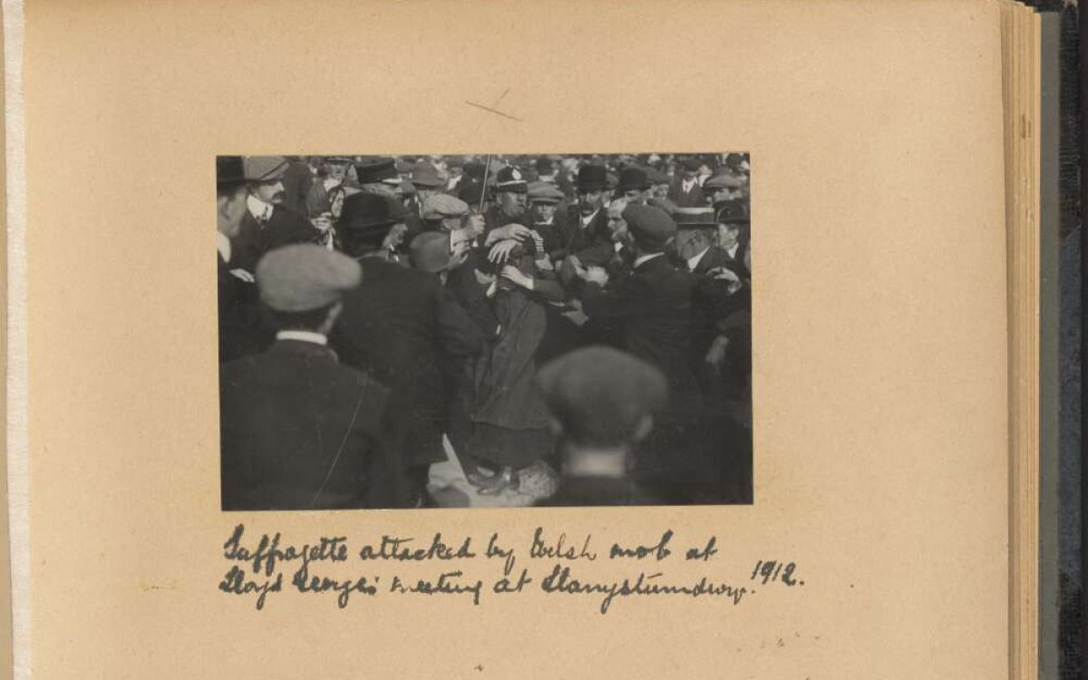 A black and white photograph in a paper album. The photo shows a group of people in a large crowd. There is handwriting below the images reading "Suffragettes attacked by [unclear] mob..."