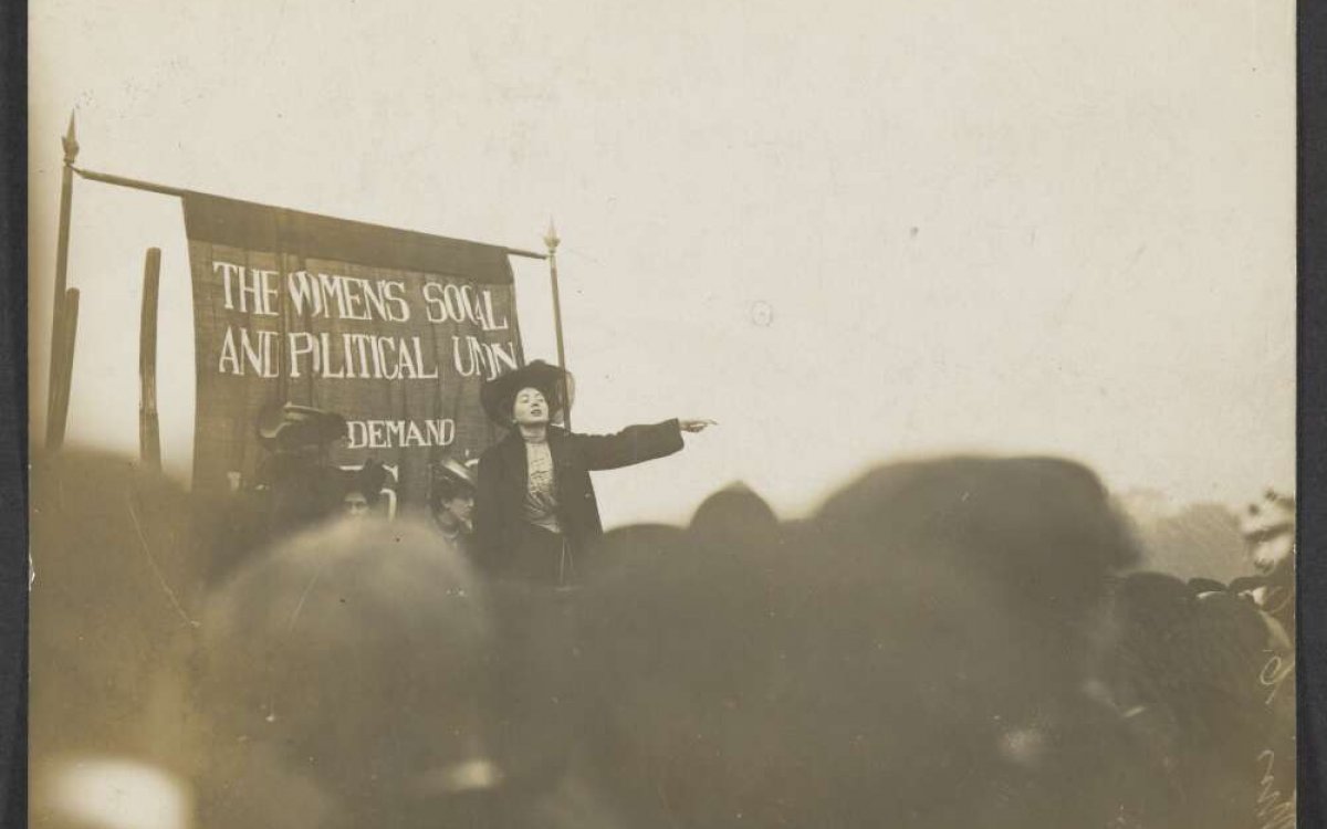 A sepia photograph of a woman wearing a black coat and hat addressing a crowd. She is standing in front of a banner that reads THE WOMENS SOCIAL AND POLITICAL UNION
