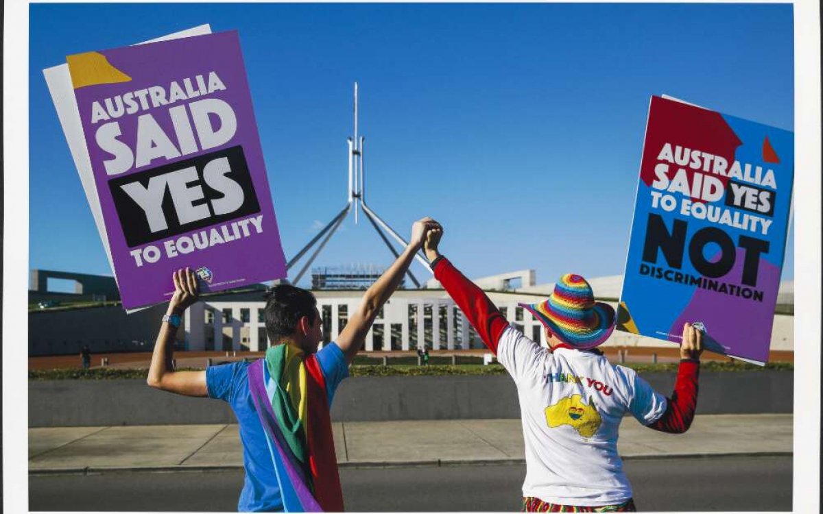 A photo of 2 people with their backs to the camera, facing Australia's Parliament House, holding hands, lifting them high. Each is holding a large sign in their outside hands that reads 'Australia said Yes to equality' and 'Australia said Yes to equality, not discrimination'.