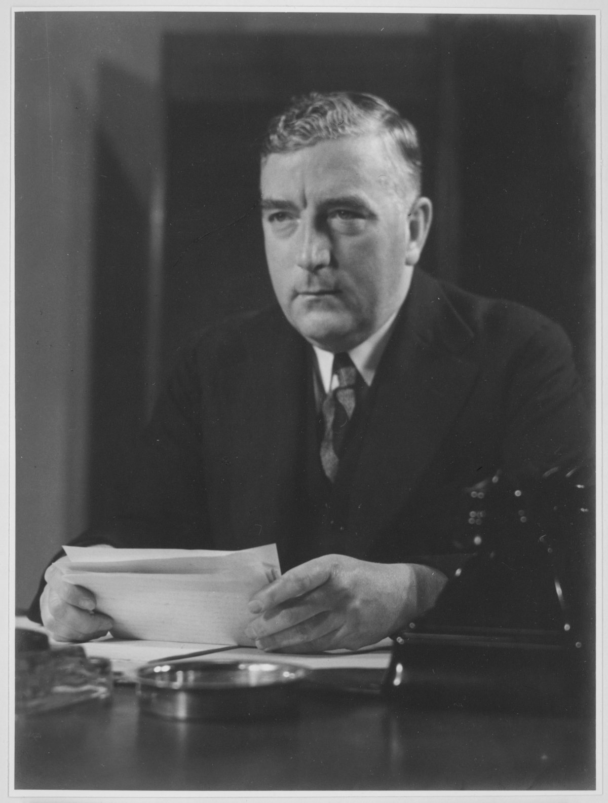Australian Prime Minister Robert Menzies sits at a desk holding several pieces of paper. He is looking melancholic and stares off camera to the left. He is a middle-aged white man with blonde hair swept to one side. He wears a dark suit and tie with a dark tie. There is a black Bakelite telephone on the table in front of him