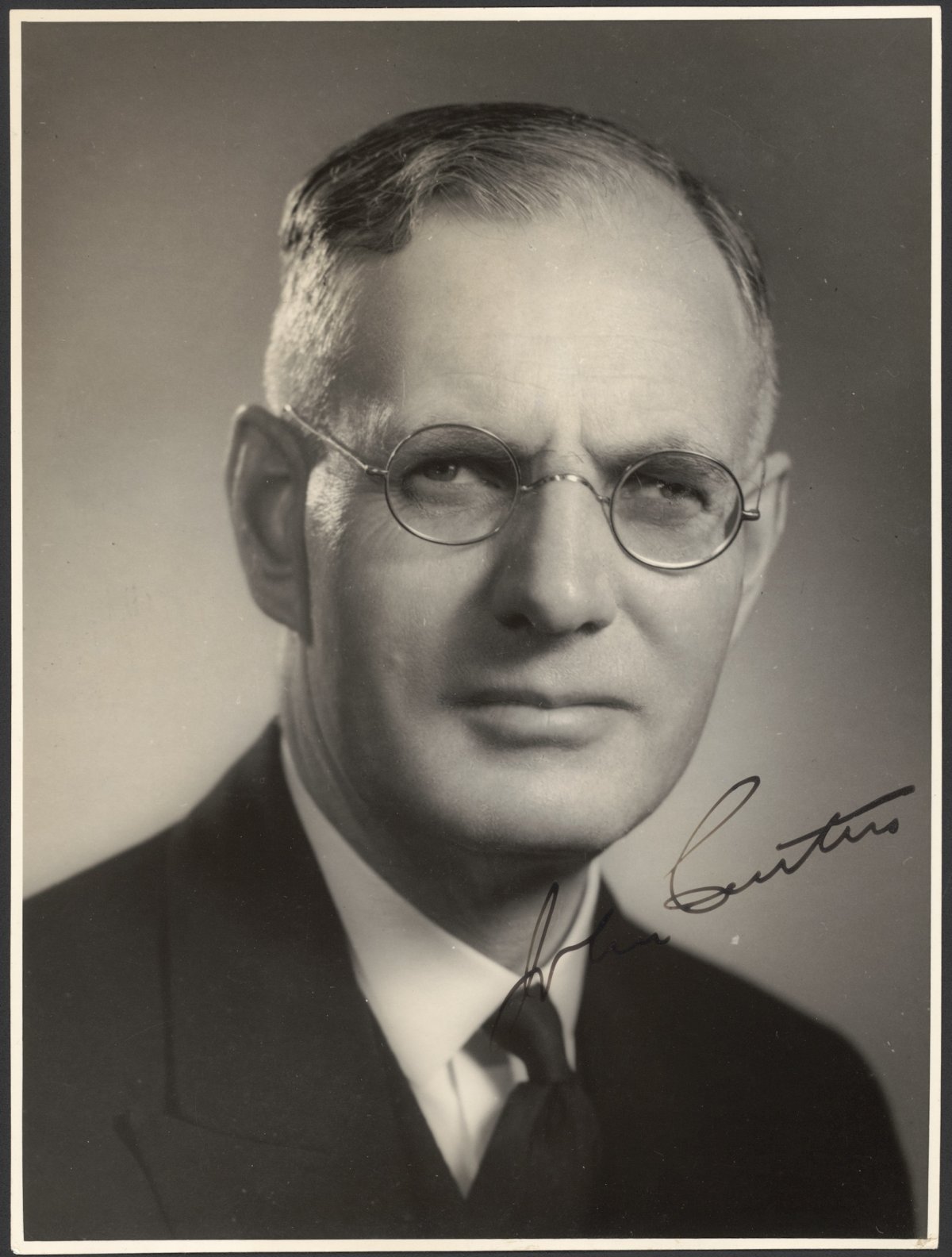 An official portrait of Australian Prime Minister John Curtain. He is a middle-aged white male wearing wire rimmed glasses. He has thinning blonde hair swept back neatly. He is wearing a dark suit and tie with a white collared shirt. He is staring directly at the camera with a stern look. In the lower right quadrant of the photograph is Curtains signature.