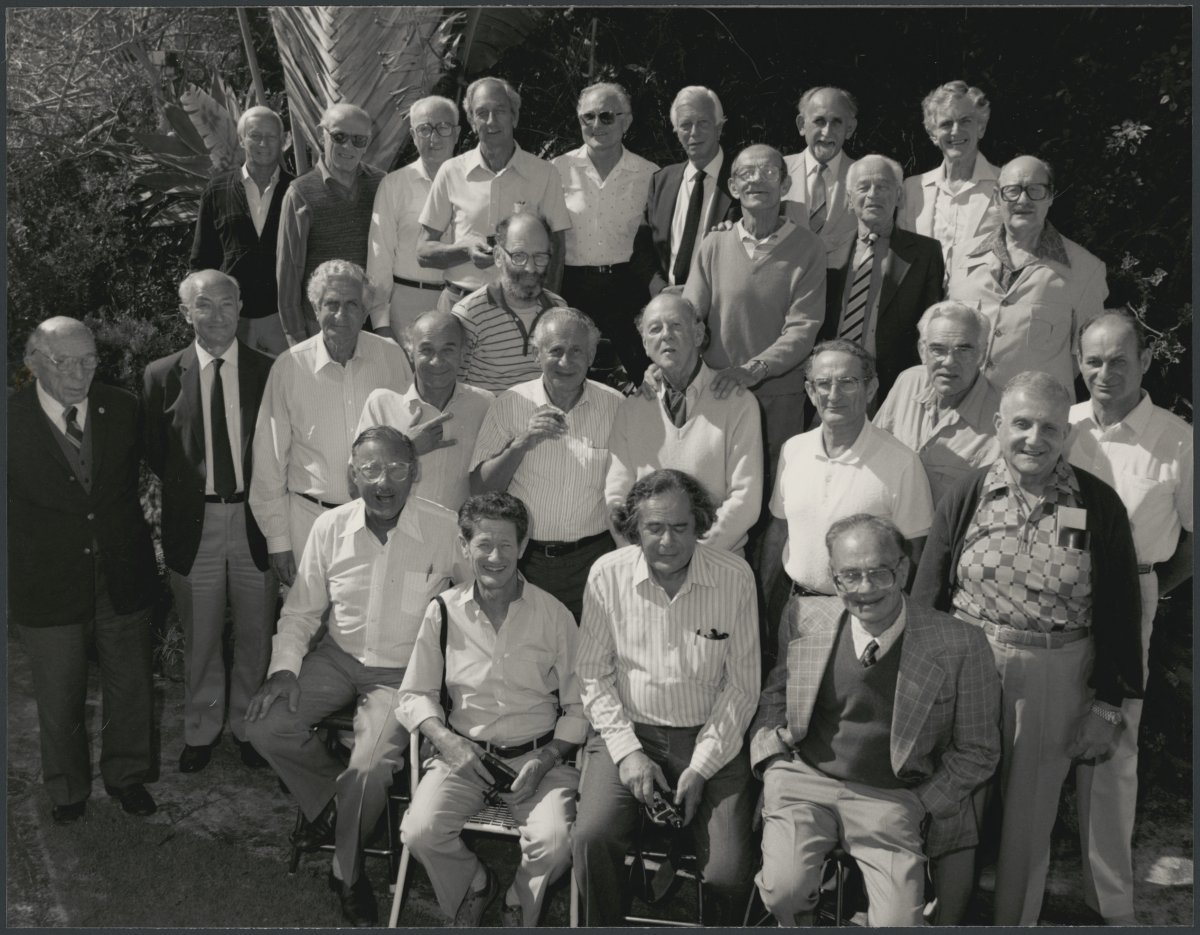 A black and white photograph of a large group of older men, posed for a group photo.  Most of the men are smiling at the camera. Some men are sitting in the front rows while others stand. They are smiling at the camera.