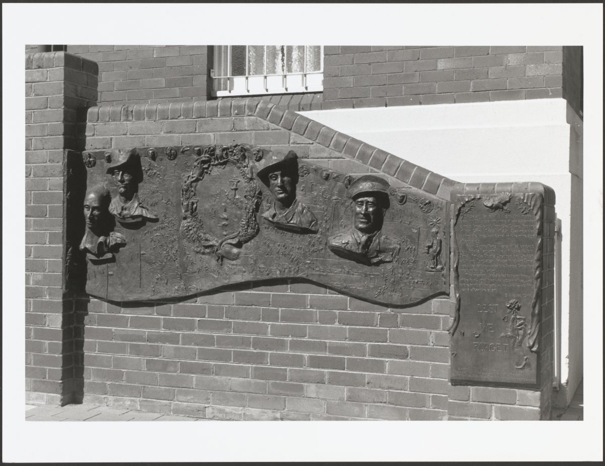 A black and white photograph of a memorial plaque. The plaque is attached to a brick wall in front of a building. The plaque features bas relief sculptures of the busts of 4 men. Two are wearing AIF slouch hats, another wears an officers flat cap and the other has no had. There is a rectangular section to the right with the words "Lest we forget"