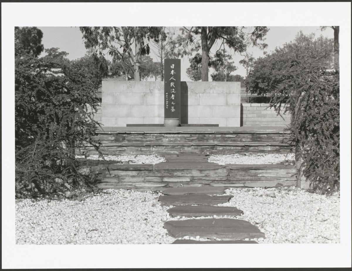 A black and white photograph of a stone memorial. In the middle of the memorial is a black stone obelisk with carved Japanese kanji. The site is surrounded by trees. There is a flagstone path surrounded by white pebbles leading up to the memorial