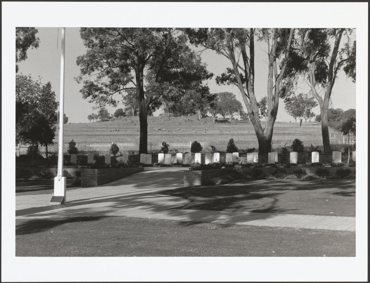 A black and white photograph of a memorial area. There are a row of 23 white headstones along the middle of the image. They are in the shade of large gumtrees. There is a white flagpole on the left of the image. In the background there is a grassy hill.