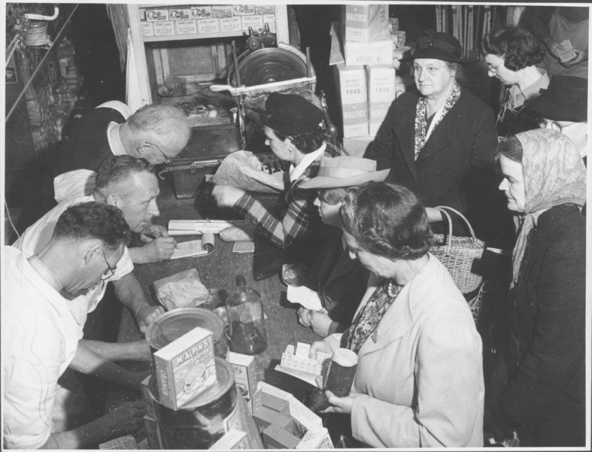 A black and white photograph showing a group of women shopping for food and supplies using wartime coupons. There are three shopkeepers behind the counter handing out goods and taking coupons