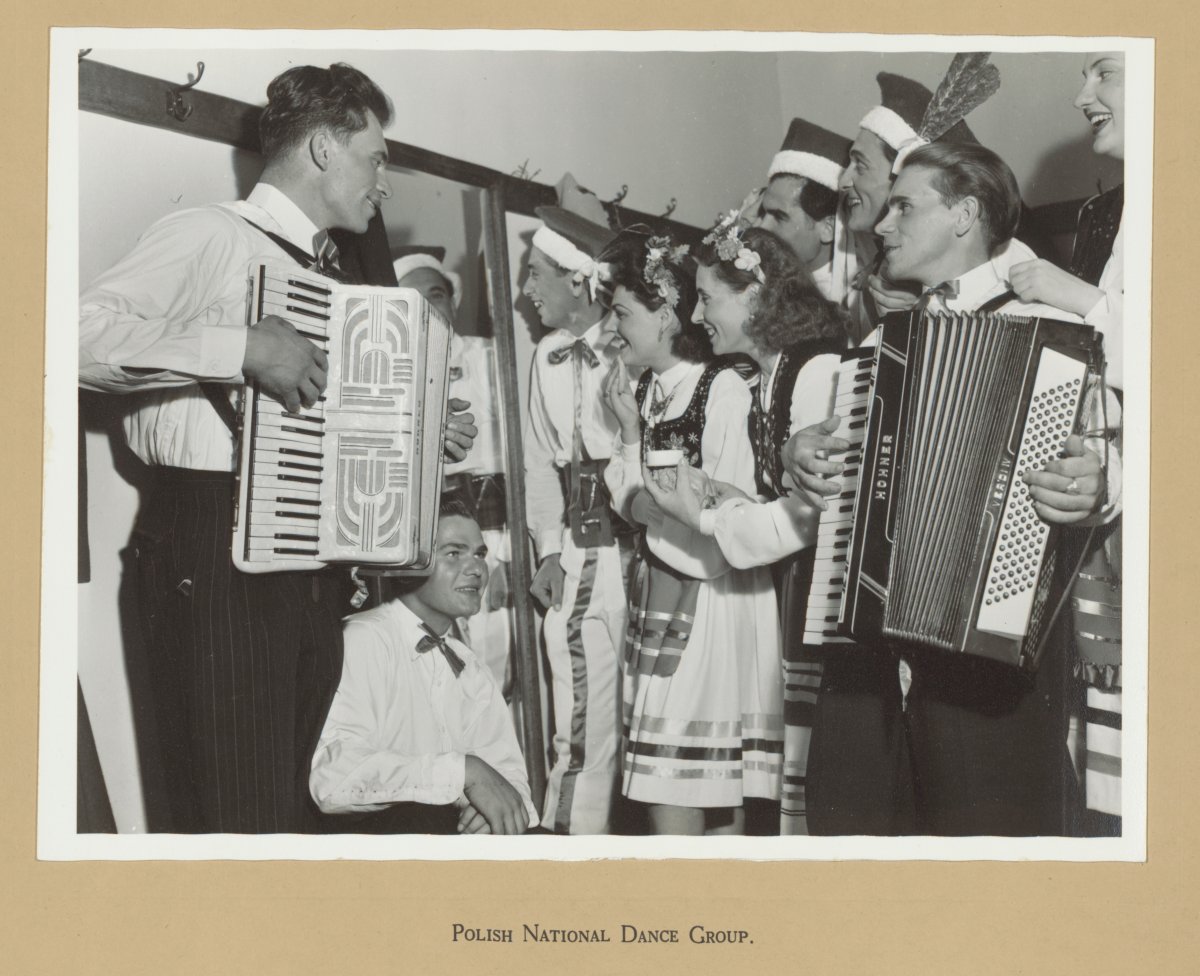 A group of people stand around two men playing button accordians. Some people are wearing Polish traditional dress others are in shirts and black pants. They look to be mid-song. Everyone is smiling.
