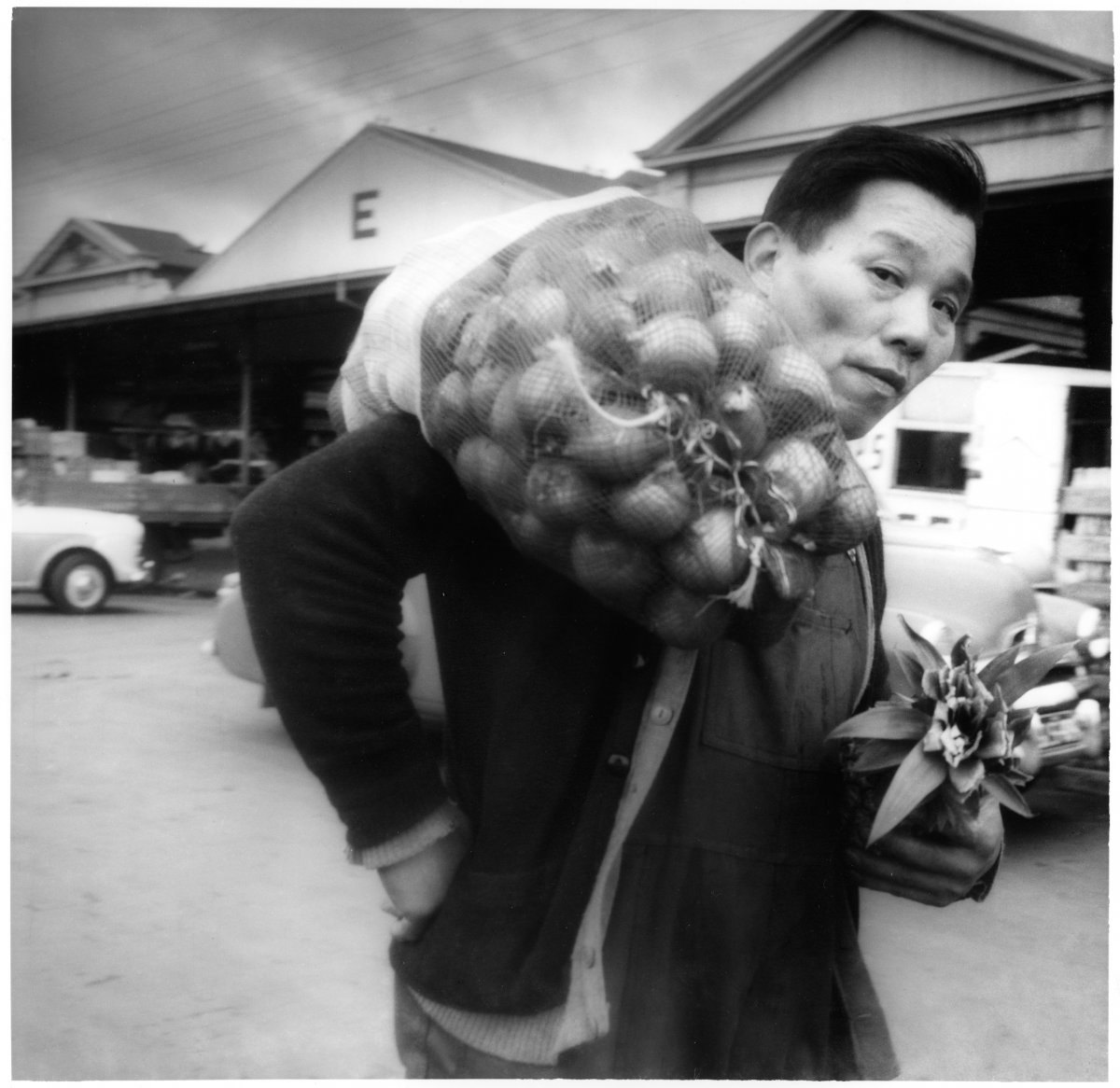 A man carriees a bag of onions over his shoulder. Behind him are the sheds of the Queen Victoria Markets, a large produce market. The man is wearing dark overalls and two cardigans