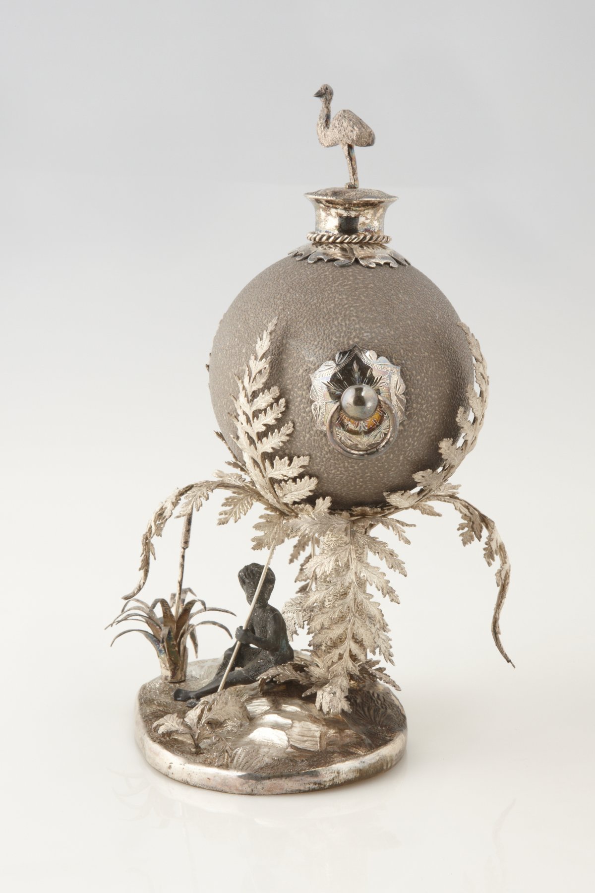 A sculpture of an emu egg on Silver-plate Stand. Underneath an Aboriginal man sits with spear. Surrounding the figure are large fern fronds and rocks.860s