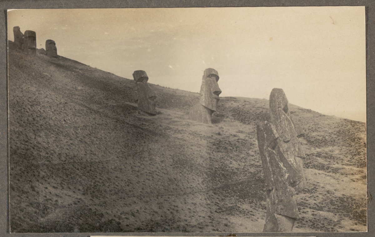 A sepia toned photograph of seven enormous stone carved heads, Moai, dotted along a barren cliff face. The statues are on various angles and have succumb to varying levels of