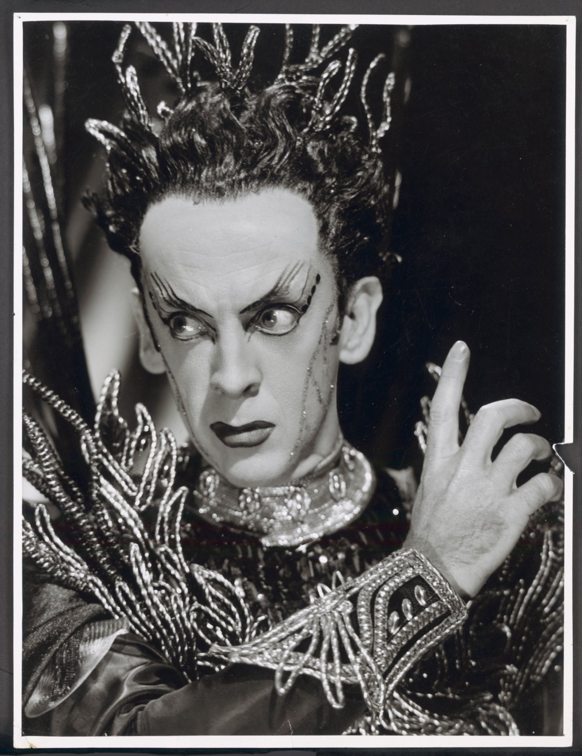 Black and white closeup photograph of Robert Helpmann in stage makeup, wearing a costume that features a beaded headpiece, cuff and collar.