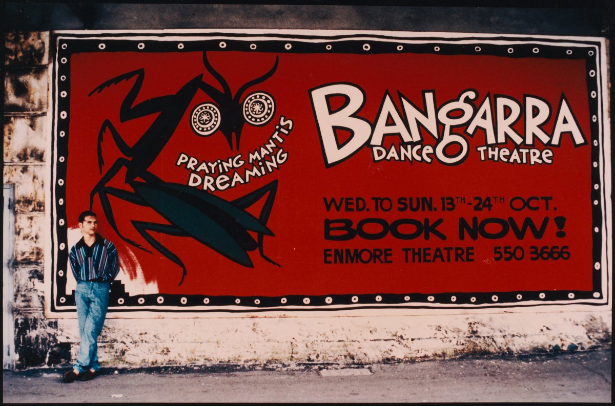 A photograph of a large billboard advertising the Bangarra Dance Theatre and the production Praying Mantis Dreaming. A large black mantis is stylised on the poster. The poster also gives the dates of the performances at the Enmore Theatre and the words BOOK NOW!. A man stands in front of the billboard. He is wearing a blue shirt and blue jeans.