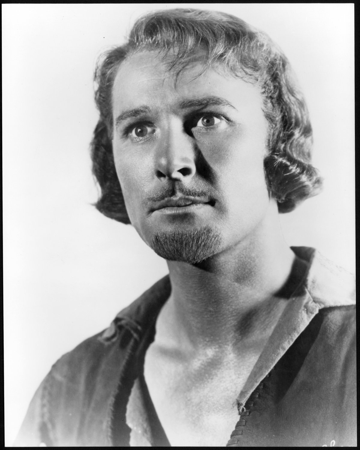 A black and white photograph of a man. He has an unbuttoned shirt showing his chest. He is staring off camera. He has a well groomed pencil moustache and a goatee. He has wavy blind hair.