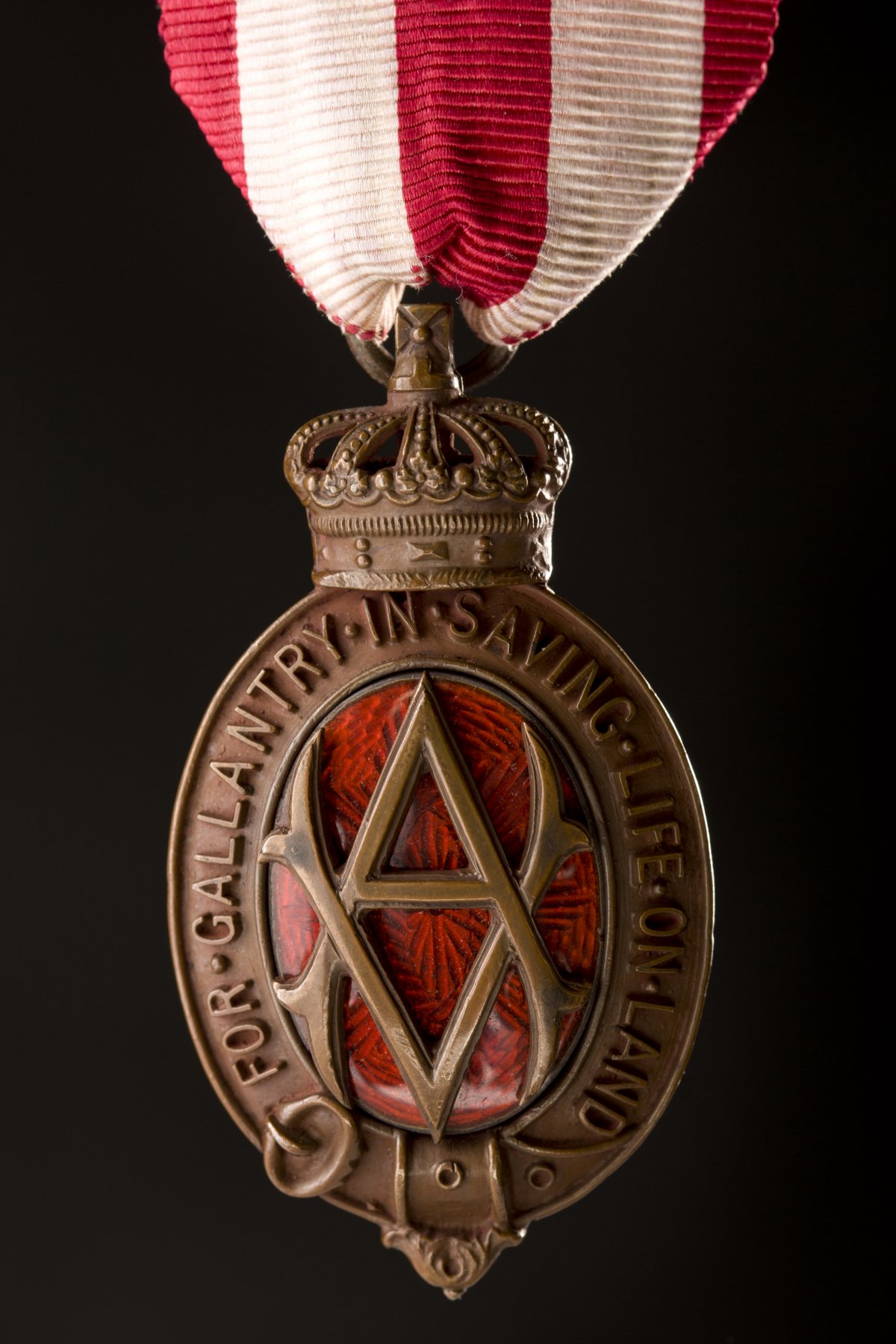 An oval shaped bronze medal. The oval is made of a belt buckled at the bottom. At the top is a stylised royal crown. In the centre of the oval is a monogram made up of an intertwined "V.A." Behind the monogram is pattered red glass. Around the oval in raised letters are the words "For gallantry in saving life on land". The medal is held by a ribbon of red and white vertical stripes 