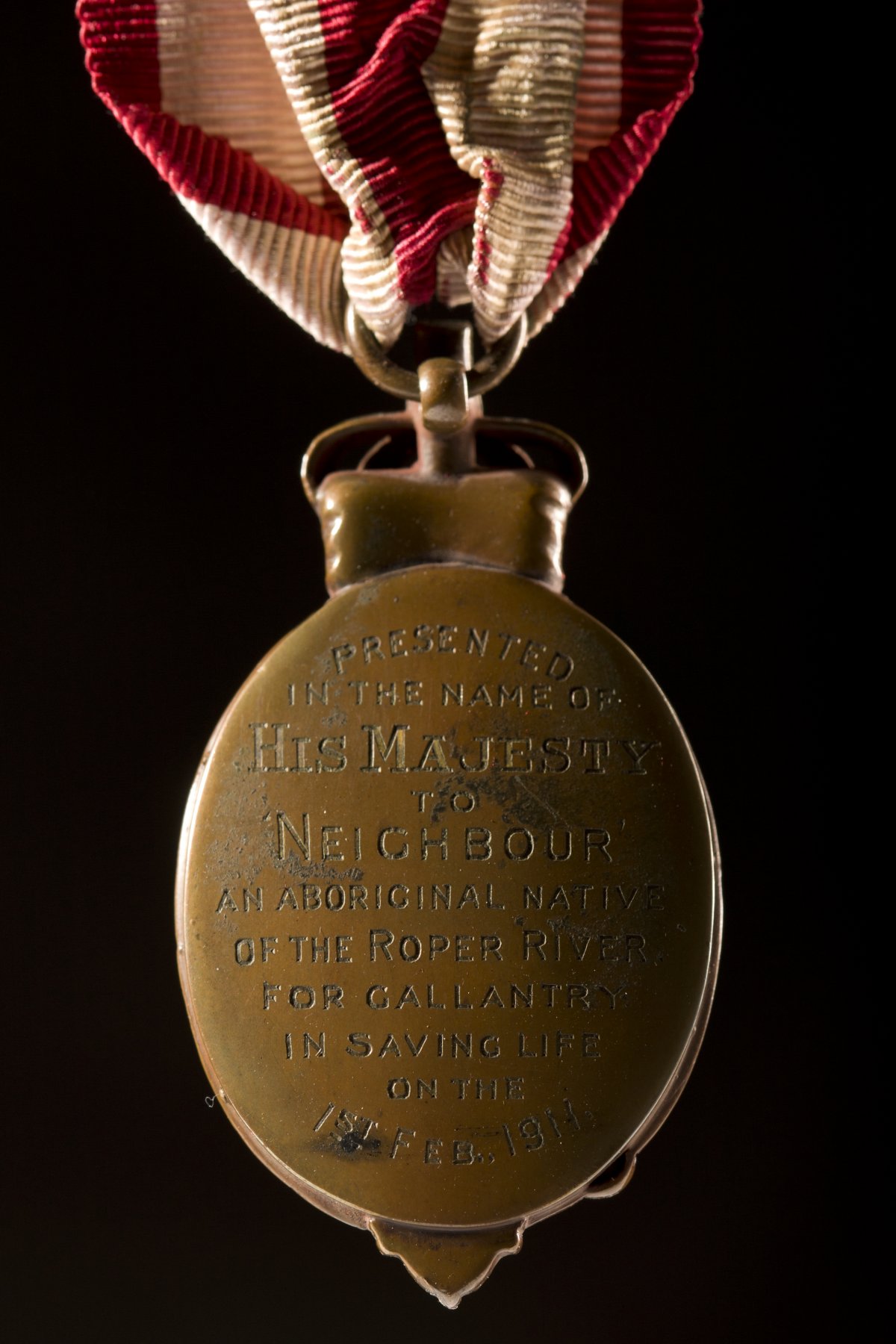 The reverse of an oval shaped bronze medal held by a red and white striped ribbon. On the back of the medal are the words " Presented in the name of His Majesty to Neighbour, an Aboriginal native of the Roper River for gallantry in saving life on the 1st Feb 1911"