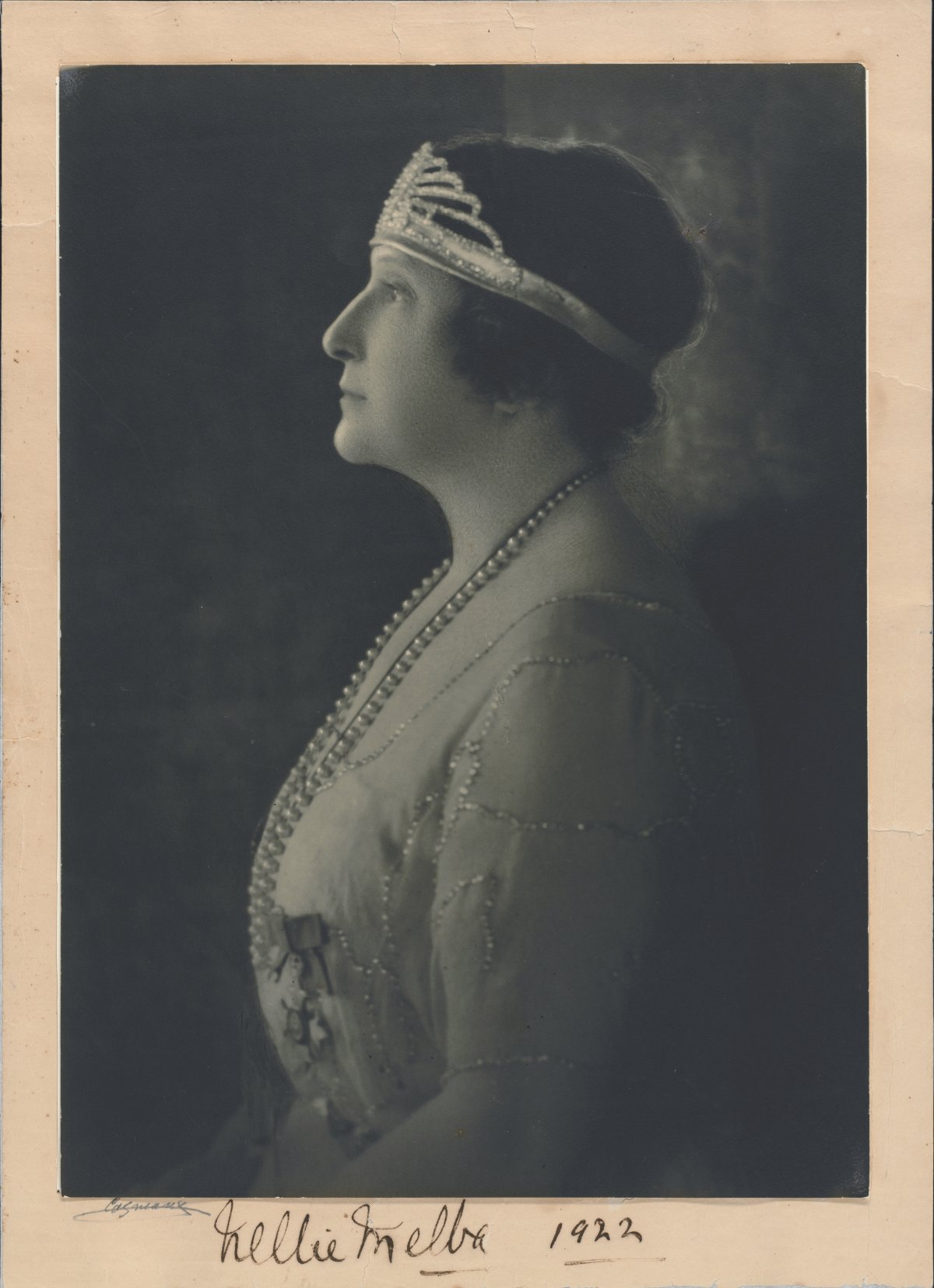 A formal portrait photograph of a lady wearing a diamond tiara. She is in profile. She has short hair and a lace gown on. She is wearing a long string of pearls. The photograph is signed "Nellie Melba 1922"