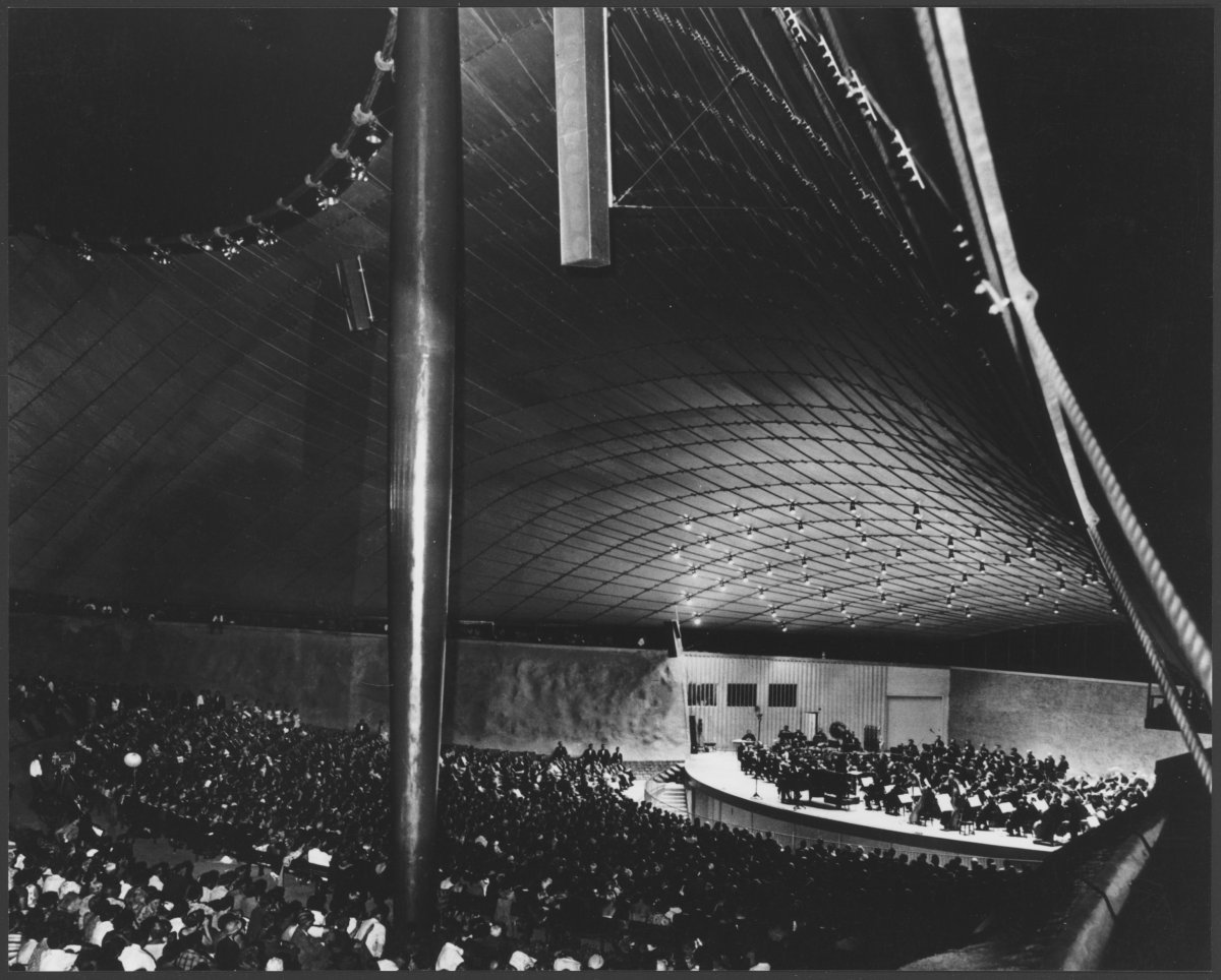 A black and white photograph of the Sidney Myer Music Bowl. A large crowd is watching a performance of an orchestra on the stage.