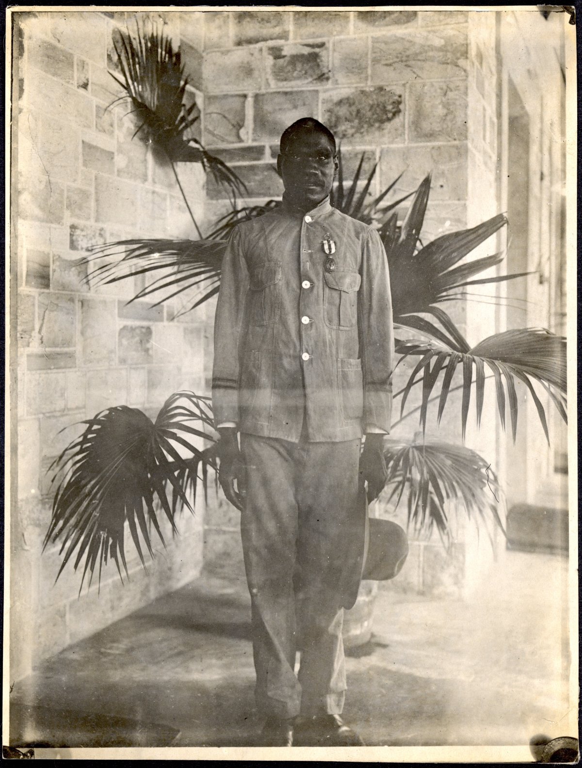A photograph of an Aboriginal man standing on the porch of a large house. Directly behind him is a large palm plant. He is staring at the camera and is standing slightly uncomfortably. He is wearing a military style tunic. Pinned to his left breast above the pocket is a medal. The ribbon is vertically striped.