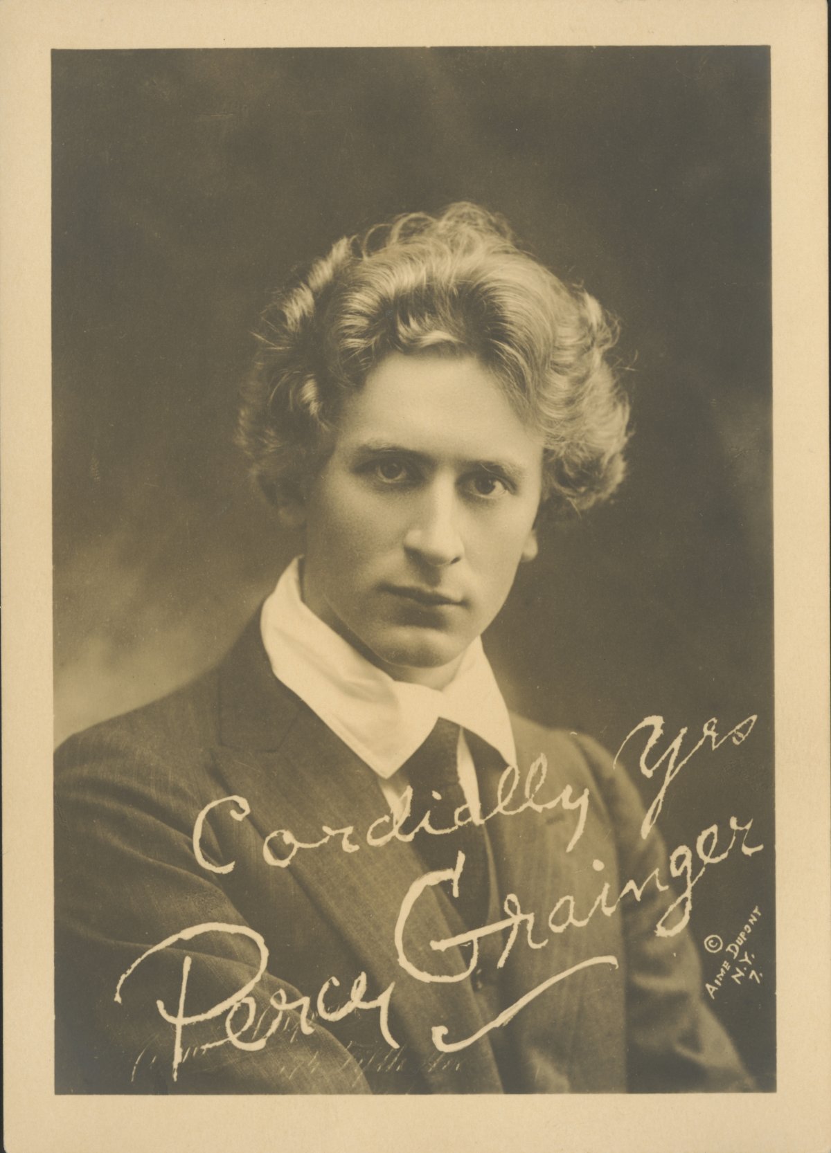 A sepia toned portrait of a young man with wavy blond hair. He is wearing a shirt with large lapels and black tie. Signed on the photo are the words "Cordially yours, Percy Grainger"