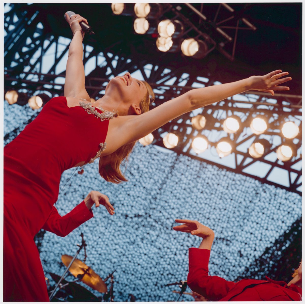 A woman in a red dress throws her hands up in the air while on stage. Behind her a backup dancer is performing. Many spotlights are shining on the stage