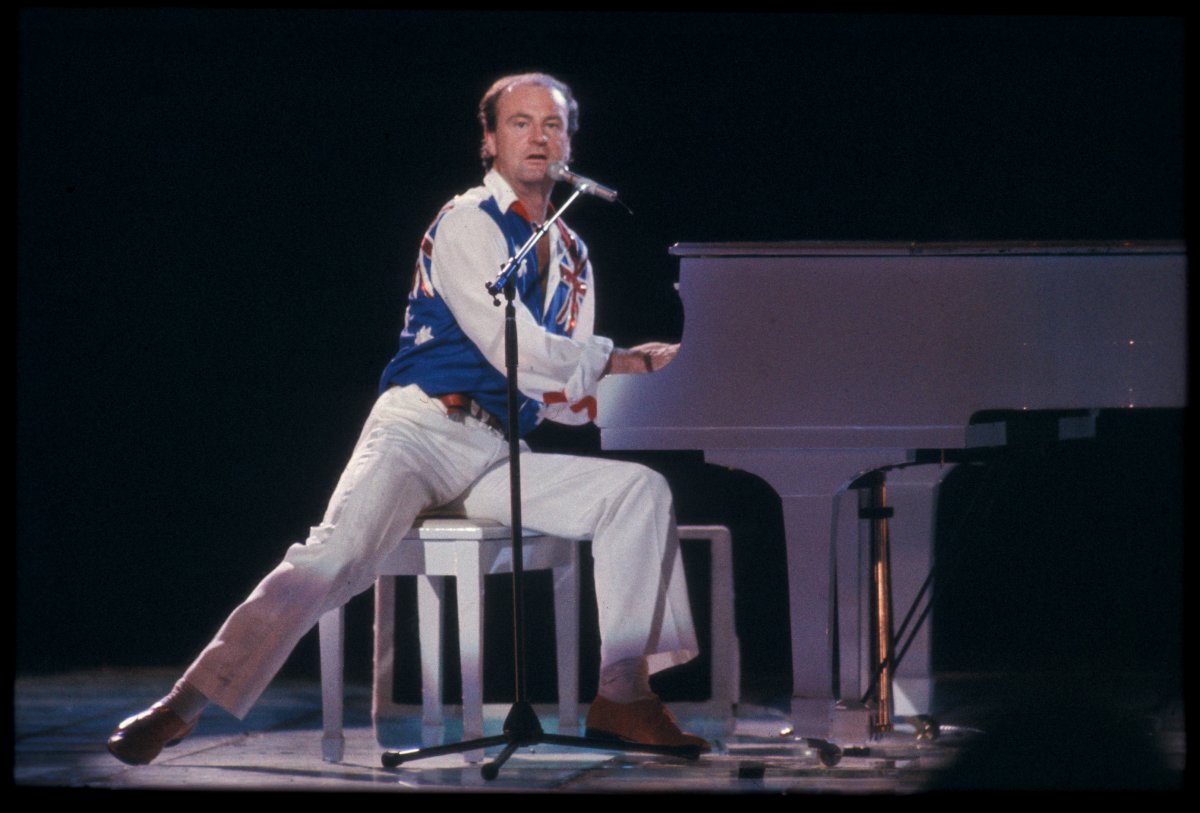 A man wearing white pants, shirt and an Australian flag vest plays a white grand piano on stage