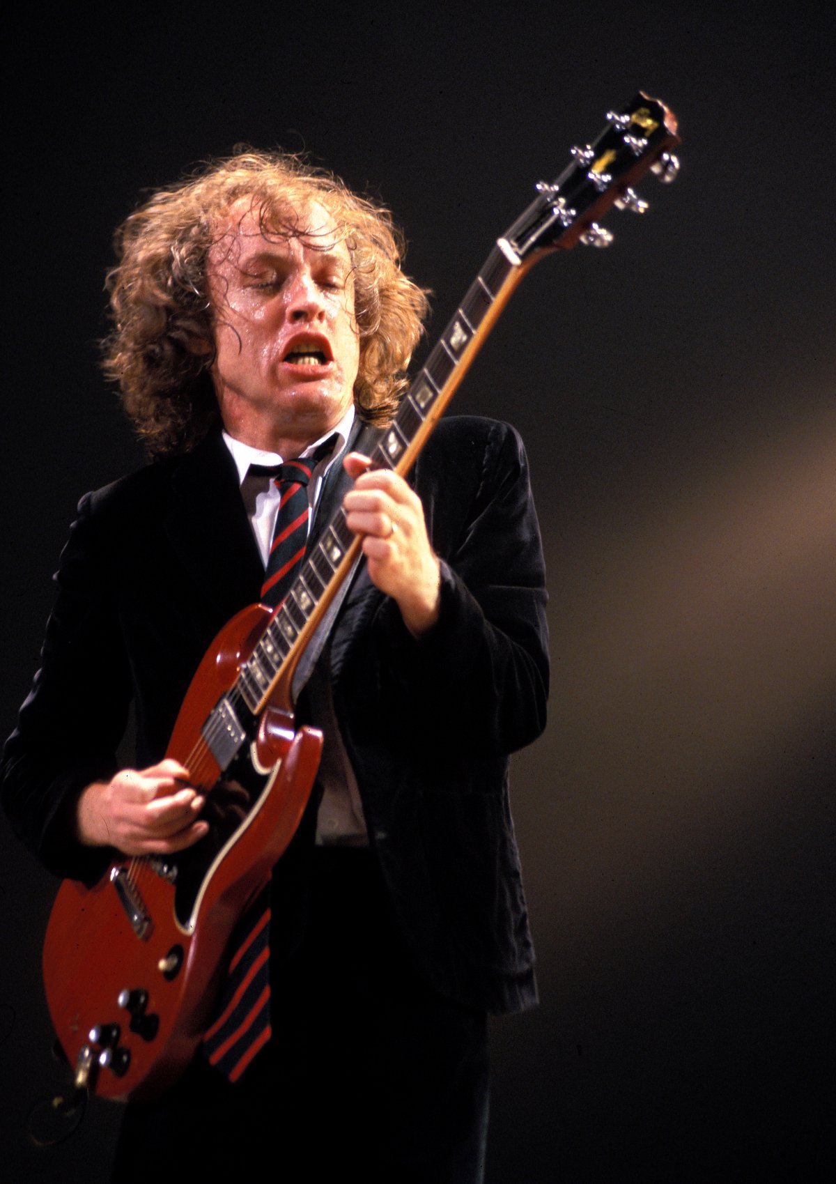 A man with long curly hair plays a guitar with great energy. He is very sweaty. The guitar is electric and has a red body. He is wearing a school uniform with a black blazer and a red and black striped tie