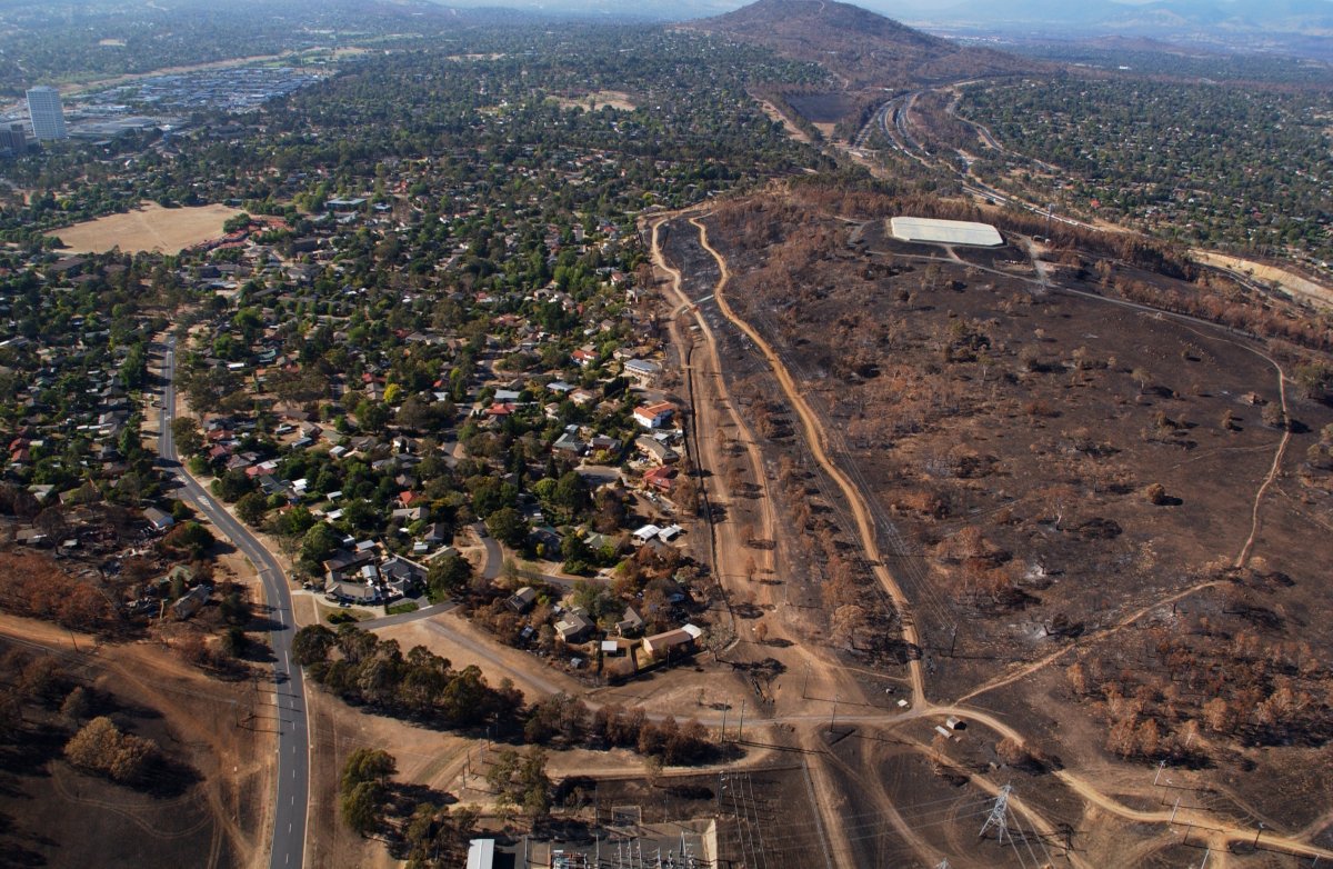 An aerial shot of a city suburb. A hill surrounding the suburb is scarred black from a large fire.