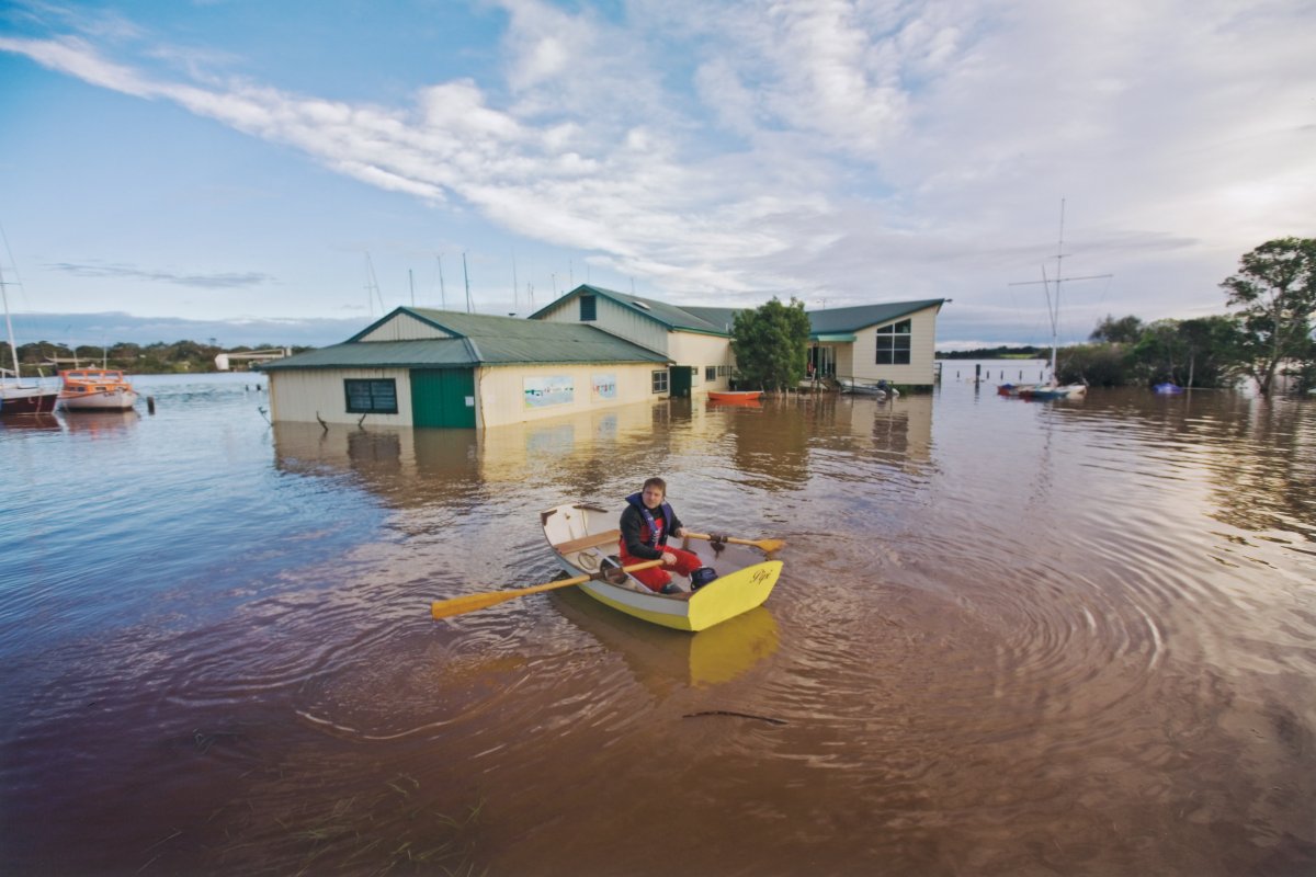 A building is partially submerged by flood water. A man in a dinghy is rowing towards the structure.