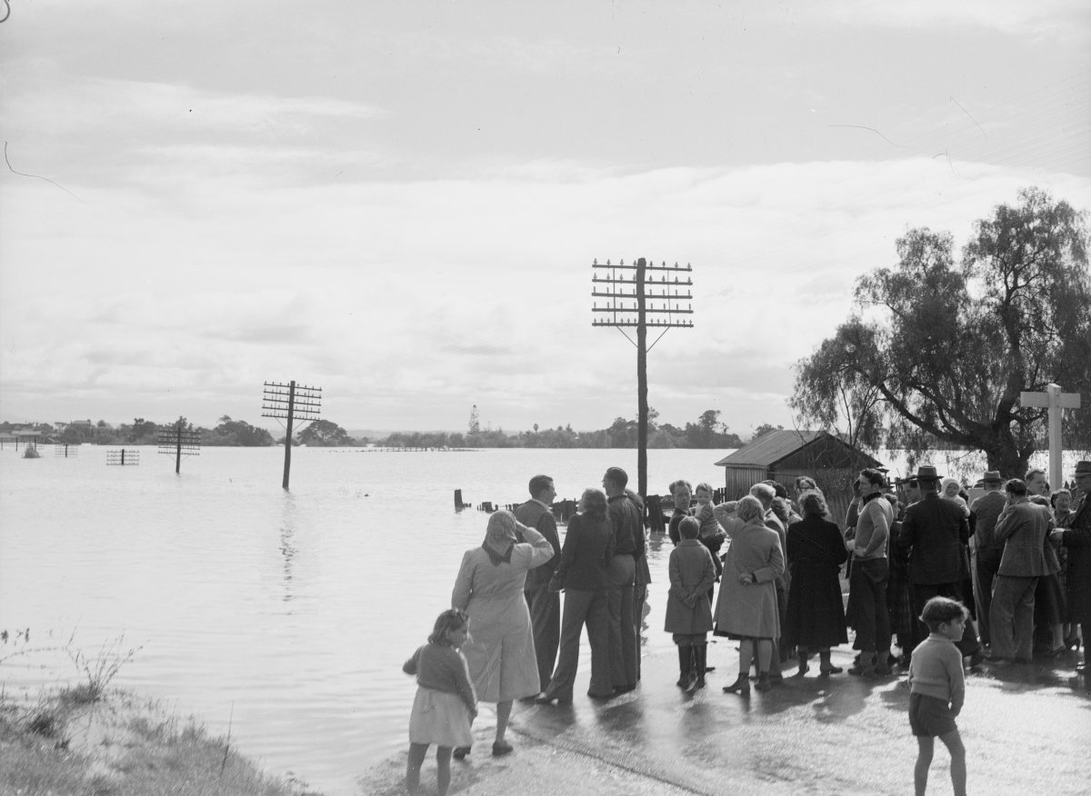 A large group of people stand on a road that is cut off by a swollen flooded river. Telegraph poles are submerged up to the head and trail off into the water.