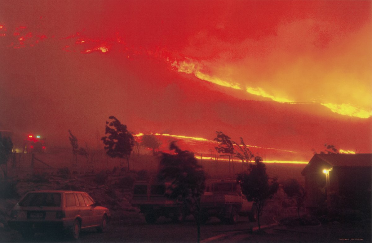 A scene of a hillside completely ablaze. A large fire front is moving up the hillside. The image is blanketed with smoke. The lights of a fire engine can be seen through the smoke. A station wagon is parked in the forground.