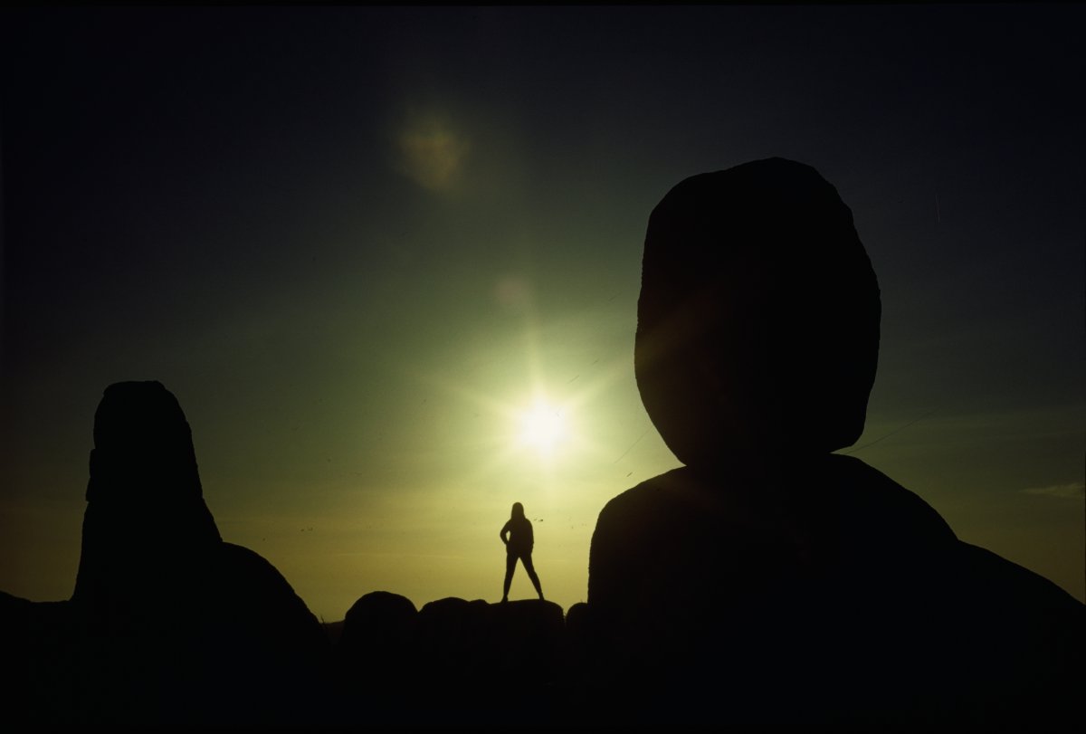 A high contrast image of two large monoliths silhouetted against a rising sun. The sky is shades of black, grey and yellow. A lone figure of a woman is silhouetted against the sun light.