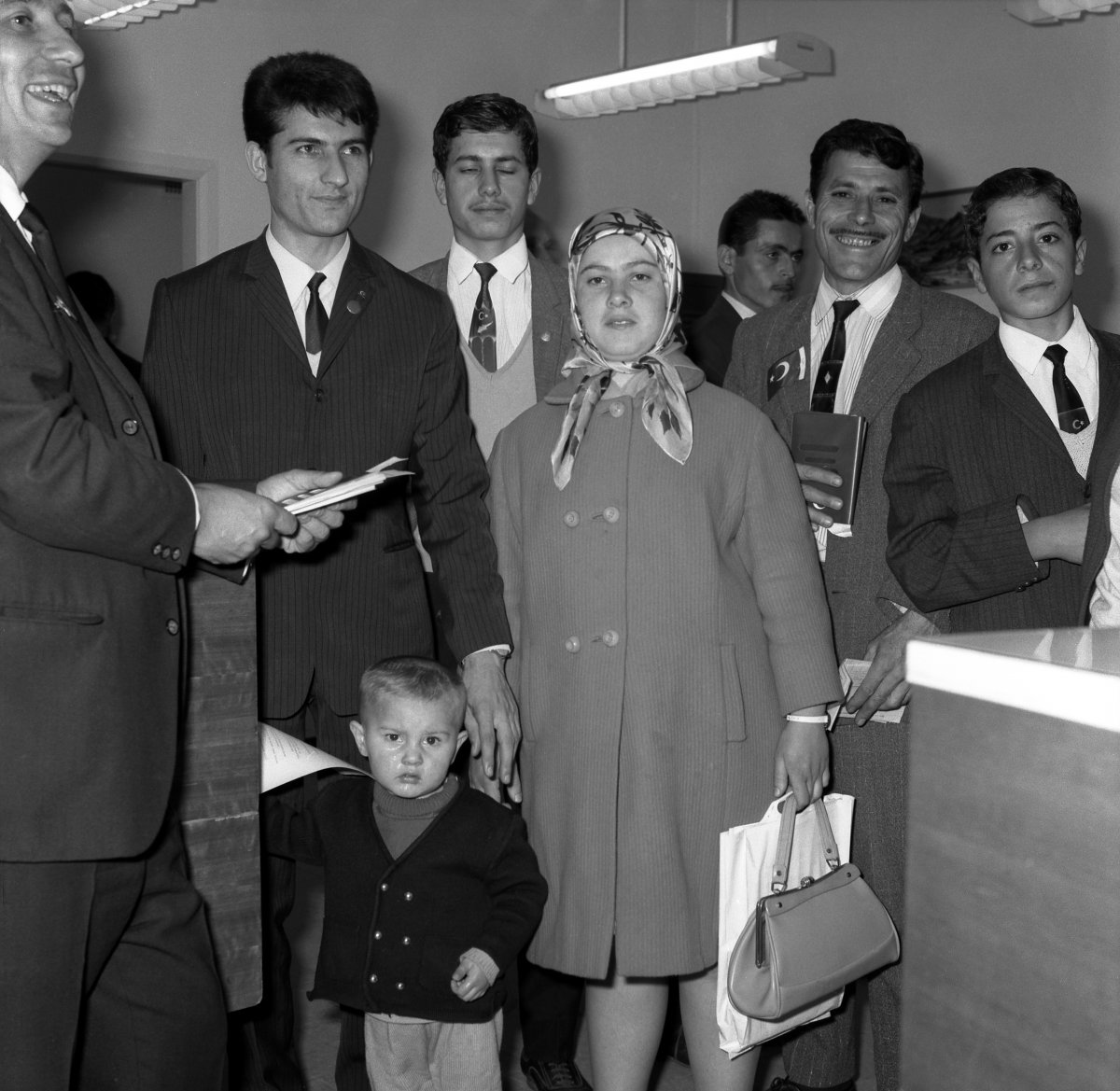 Eight people stand infront of a desk. There are 6 men, one woman and a small boy. The woman is wearing a headscarf and carrying a shopping back and a handbag. One man is holding tickets/papers. Another man holds a passport.