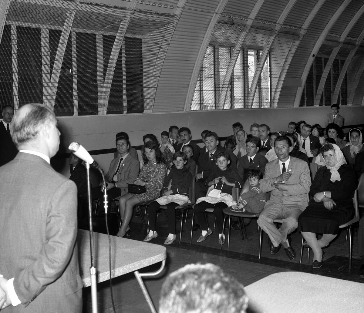 A photograph of a large group of people seated in an auditorium. A balding man is standing at a microphone addressing the audience.