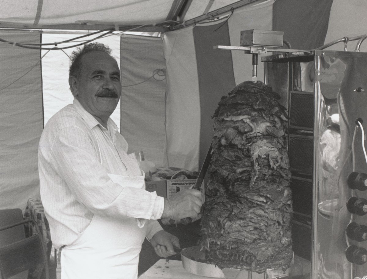 A man with a dark moustache and a white apron is carving meat from a revolving pile to be used for kebabs. He is inside a tent.