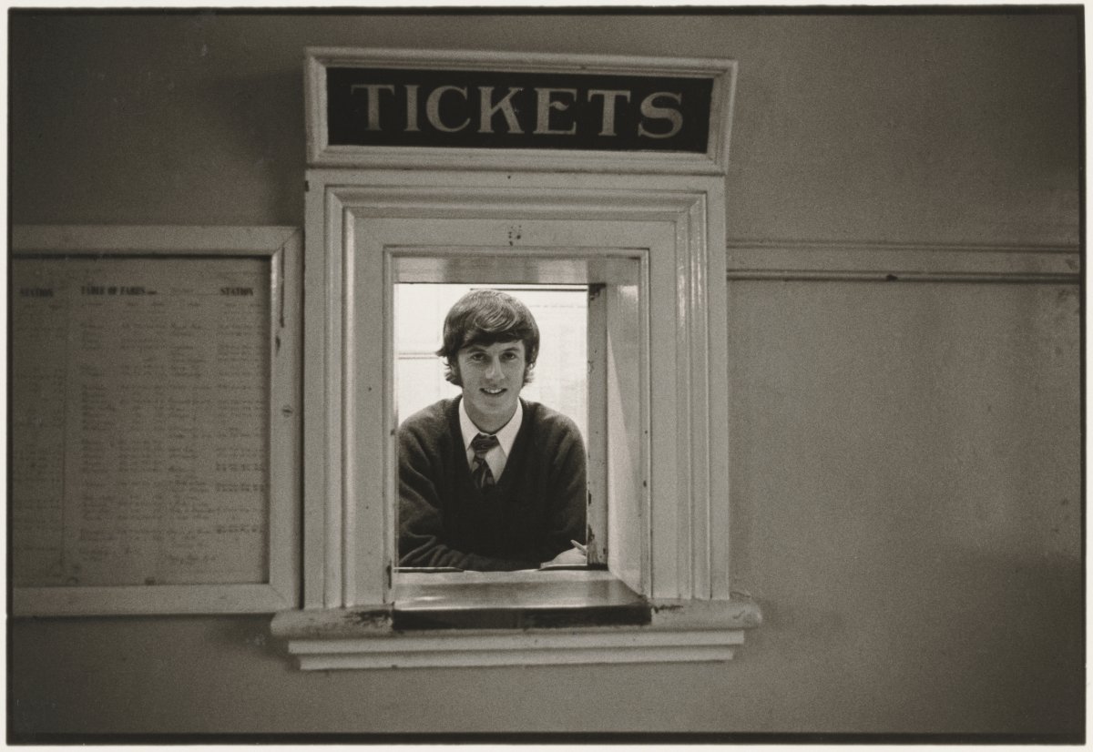 A young man leaning on the ledge of a ticket booth with a program sitting to his left