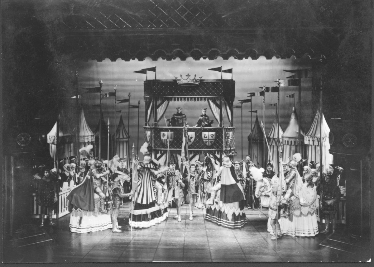 A black and white photograph of a theatre performance. The actors are dressed as medieval knights and soldiers. They are looking upstage at two figures in a royal box.
