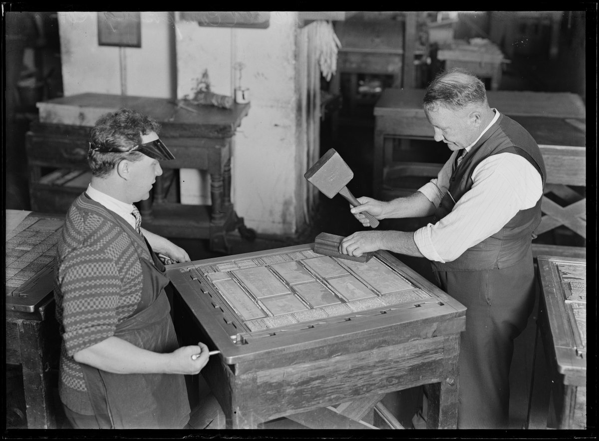 A black and white image of two men setting a page up with typeface. One man is setting the screws of the frame while another man makes  sure all the letters are set by hitting them with a wooden mallet.
