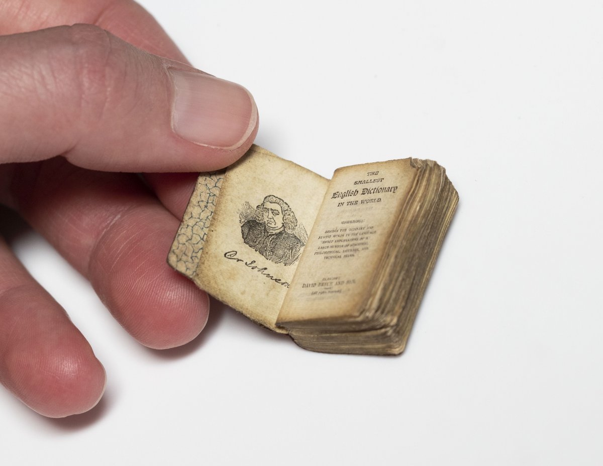 A rare miniature book opened to the title page. An illustration of a person on the right, text on the left. Someone holds the book open. The book is about the side of a large thumb nail.