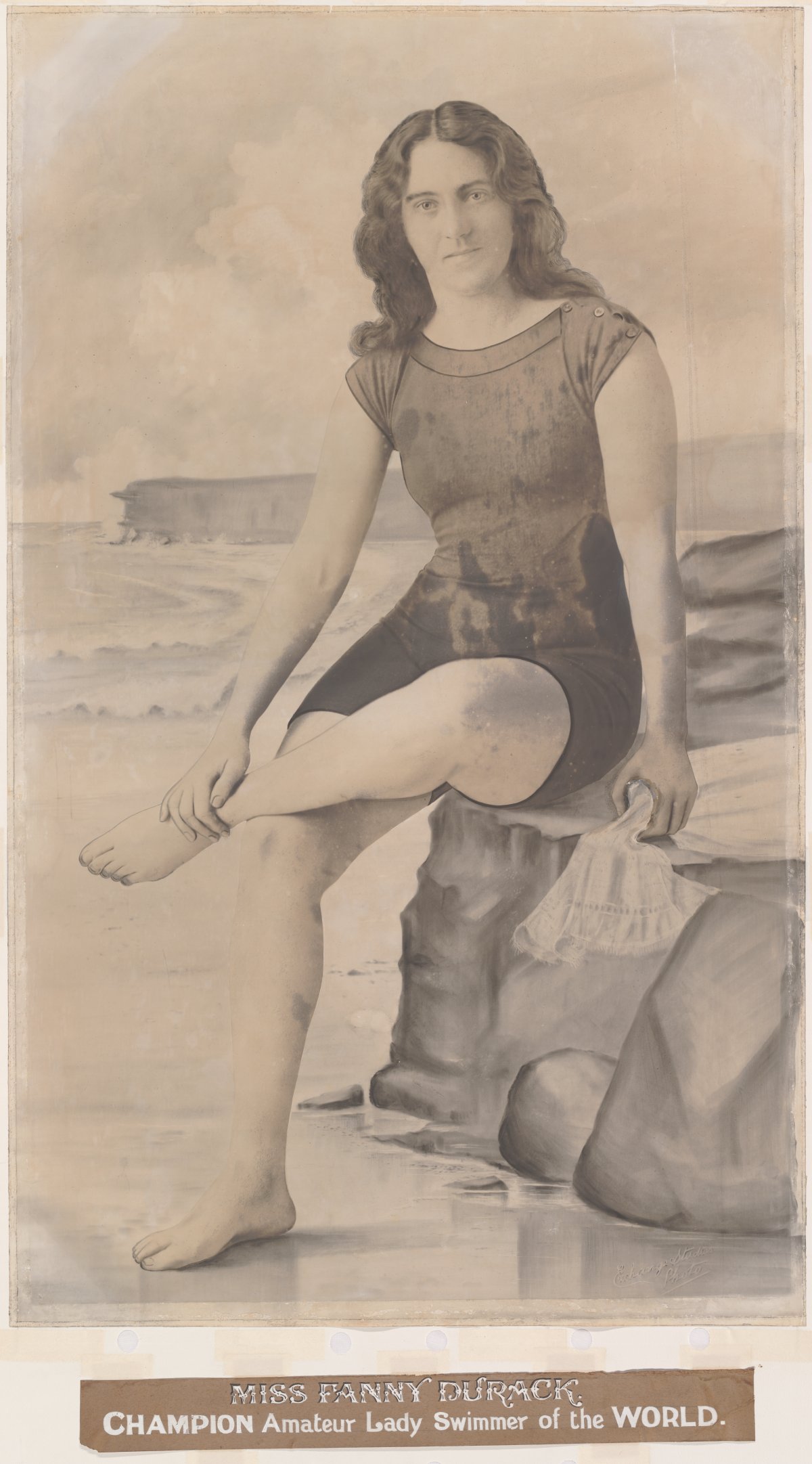 A sepia toned photograph of a woman sitting on a rock. She is wearing an old fashioned swim suit. She is sitting crossed legged. Beneath her is a caption saying ""MISS FANNY DURACK" Champion Amateur Lady Swimmer of the World"
