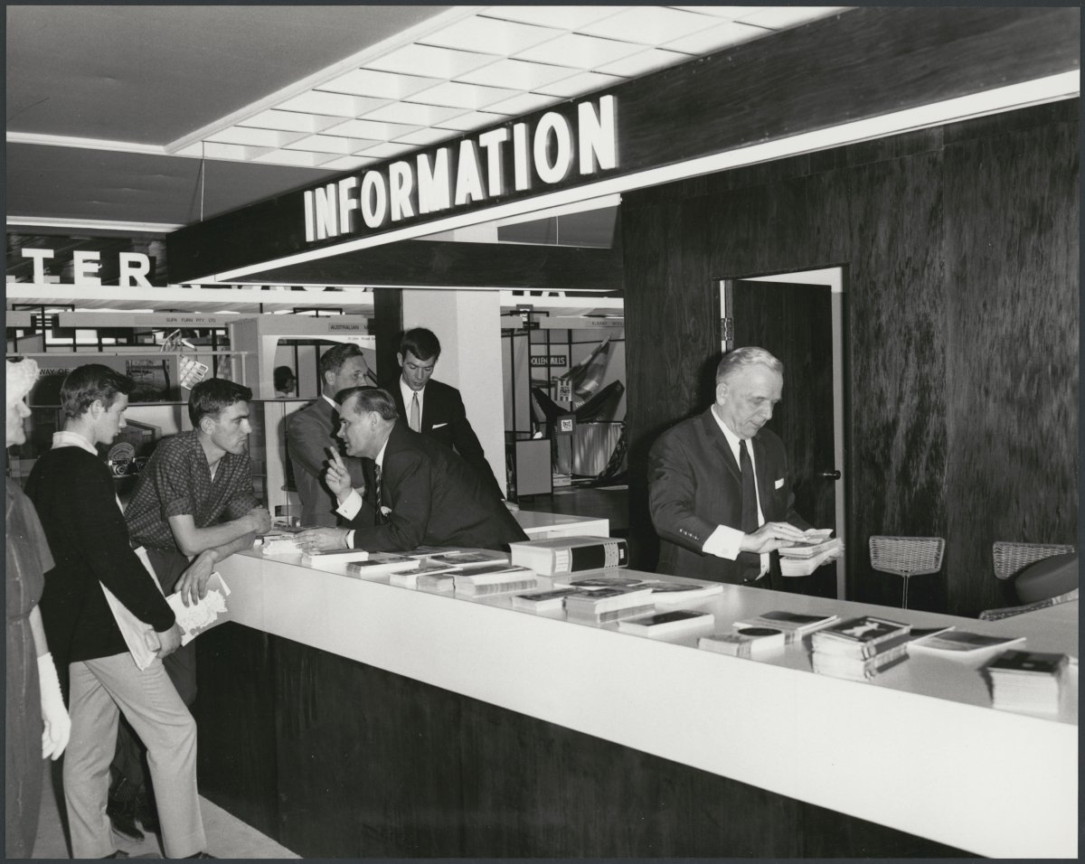 A black and white photograph of a group of men standing at a white information desk. Above the desk is a large sign that says INFORMATION. Men behind the counter and speaking and hanging out brochures.