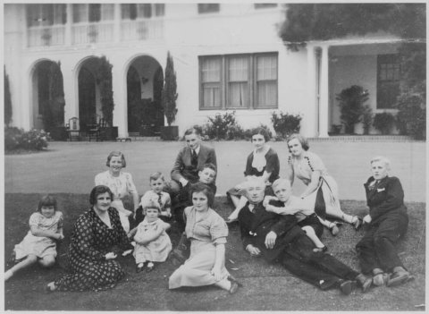 Black and white photograph features a number of people and small children lounging on lawn outside The Lodge in Canberra