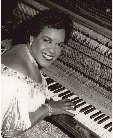 A woman playing piano is looking back at the camera, smiling. She is wearing a white dress as well as a necklace, earrings and bracelet.