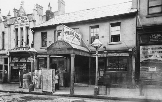 Black and white photograph of a row of shopfronts. One facade reads 'Theatre Royal'.