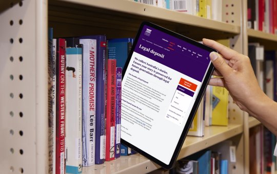 Tablet being pulled from a bookshelf like it's a book. On the screen is a page from the National Library's website sharing information about 'Legal Deposit'